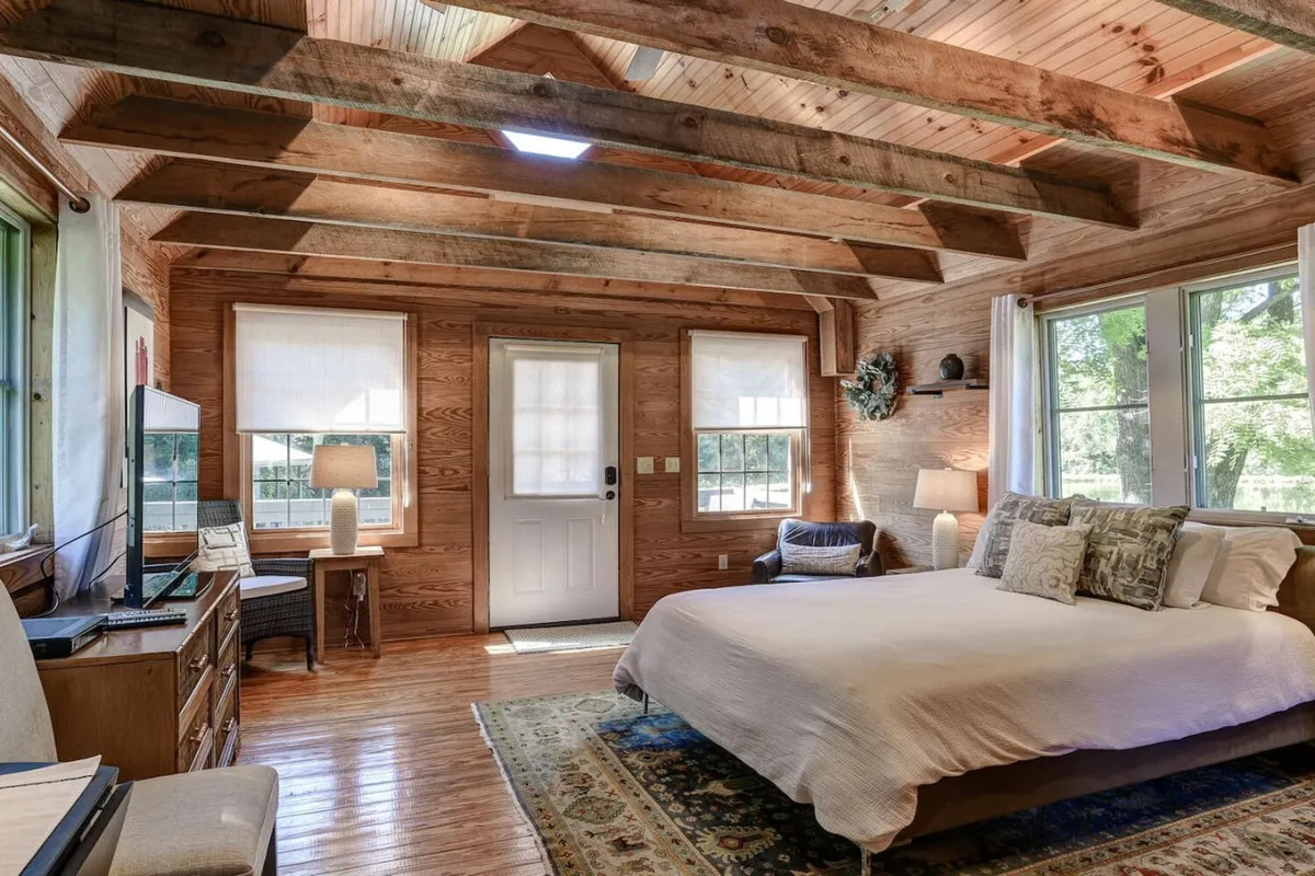 A tranquil bedroom scene with a cozy bed, large windows allowing natural light, a bedside lamp, and a soft rug. The space feels intimate and ideal for relaxation or reading.