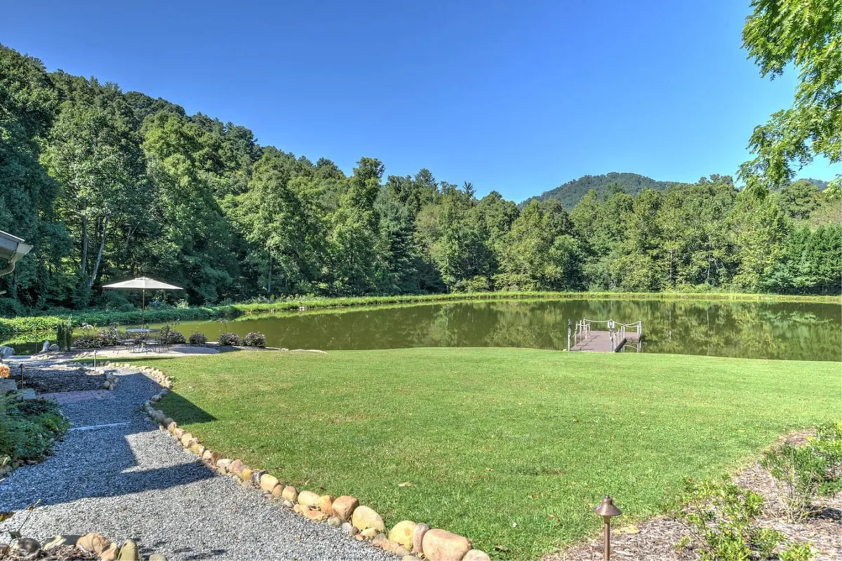 A peaceful lakeside scene featuring a calm lake, lush greenery, a dock extending over the water, and a pathway leading to the shore. The setting is tranquil, ideal for fishing, nature walks, or quiet moments with the group.
