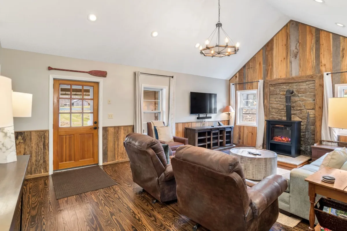 A cozy living room featuring a prominent fireplace, wooden walls, and comfortable seating arranged for conversation. Natural light streams in through windows near the front door, illuminating the rich wood floors and making the space feel warm and inviting. The scene suggests a space where people can relax, socialize, or enjoy a quiet moment by the fire.
