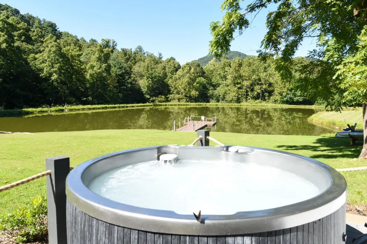 A serene exterior setting with a hot tub overlooking a tranquil lake, a dock, and surrounding mountains and trees. The hot tub is poised for relaxation, while the lake and dock invite fishing, picnicking, or simply unwinding in nature.