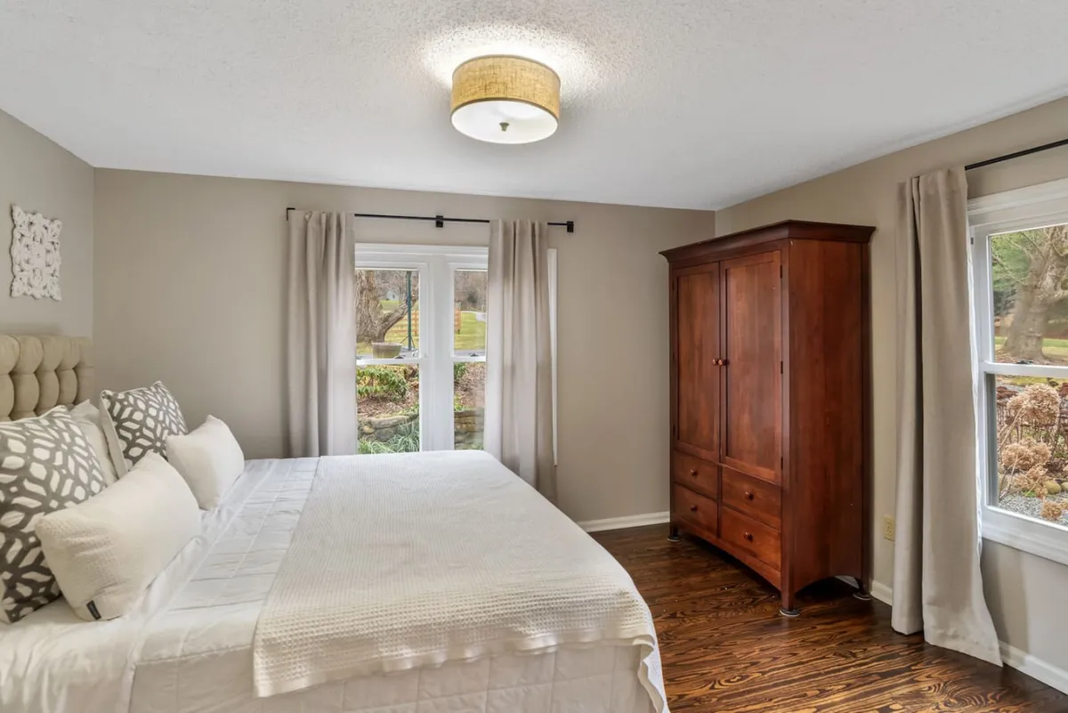 A tranquil bedroom with a bed dressed in soft linens, a wooden wardrobe, and windows that let in natural light. The view through the windows suggests a gentle connection to the outdoors, creating a perfect spot for reading or contemplation.