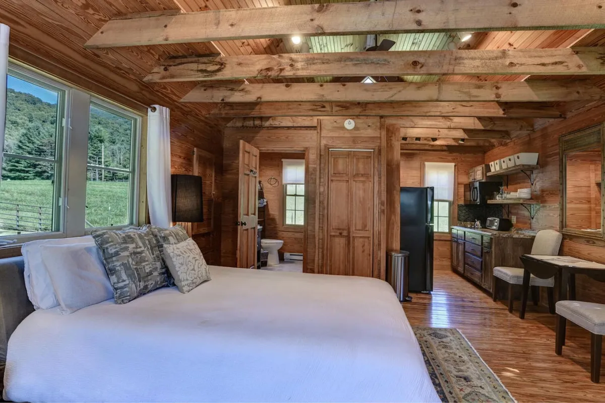 The guest cottage interior includes a comfortable bed with decorative pillows, exposed wooden beams, a kitchenette, and large windows offering scenic views. The space is tranquil and versatile, perfect for privacy and relaxation.