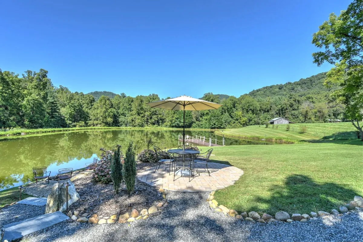 This image features a tranquil pond bordered by a lush tree line, a patio area with an umbrella, and distant mountains. The peaceful water setting offers a sense of private retreat and the patio invites outdoor dining or quiet relaxation beside nature.