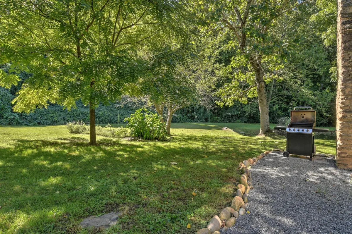 A welcoming exterior scene showcases a grill on a grassy lawn surrounded by trees, with a stone pathway leading to the space. The setting evokes summer barbecues, picnics, and the freedom for children to play on the grass. The natural landscape enhances the outdoor, family-friendly atmosphere.
