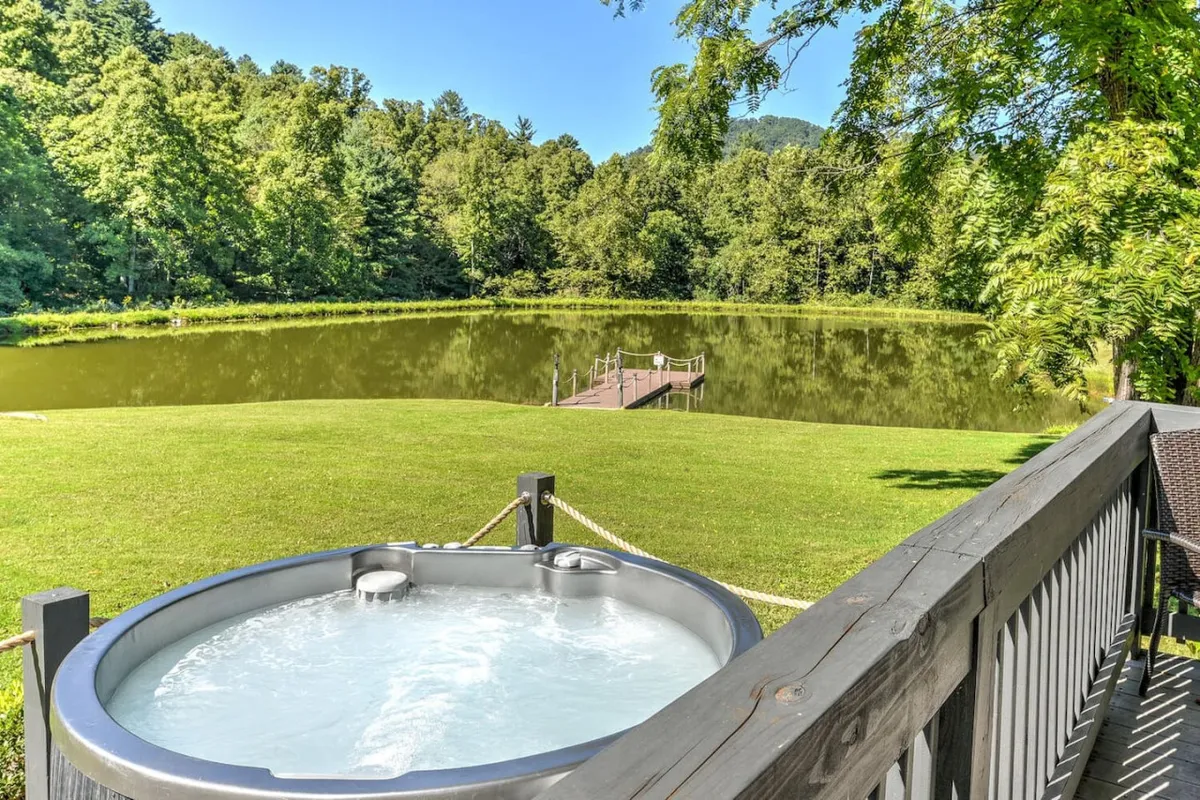 A modern hot tub is nestled on the edge of a calm lake, surrounded by vibrant greenery and a wooden dock. The setting is open to the sky, offering an inviting spot to relax, unwind, and stargaze after a day’s adventures.