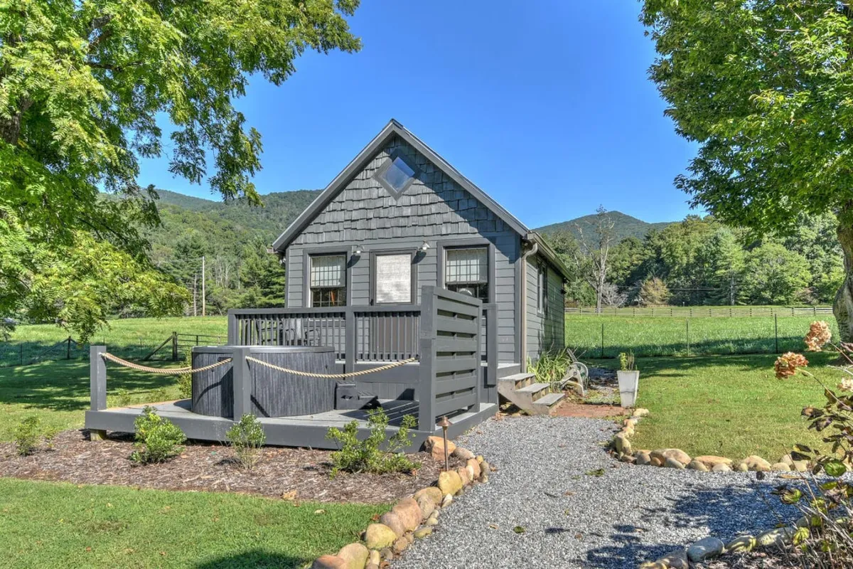 A charming gray cottage sits amid lush green surroundings, with a wooden deck offering a spot to relax and soak in the sweeping mountain backdrop. The scene radiates tranquility and seclusion, evoking the perfect countryside escape ideal for couples seeking privacy and romantic moments.