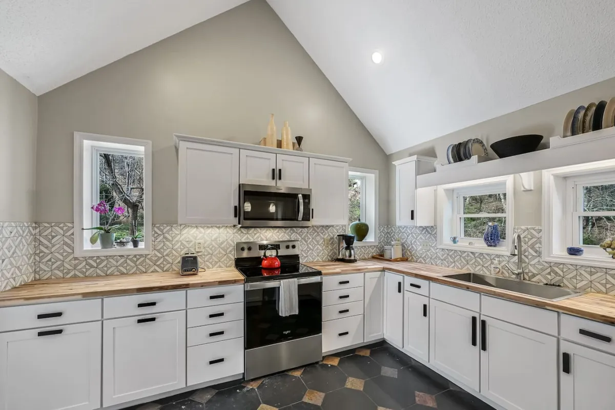 A warm, welcoming kitchen bathed in natural light, featuring stainless steel appliances, wooden countertops, a geometric tile backsplash, and visible windows that likely offer scenic farm views. The arrangement suggests ample counter space and a harmonious blend of rustic and modern design.