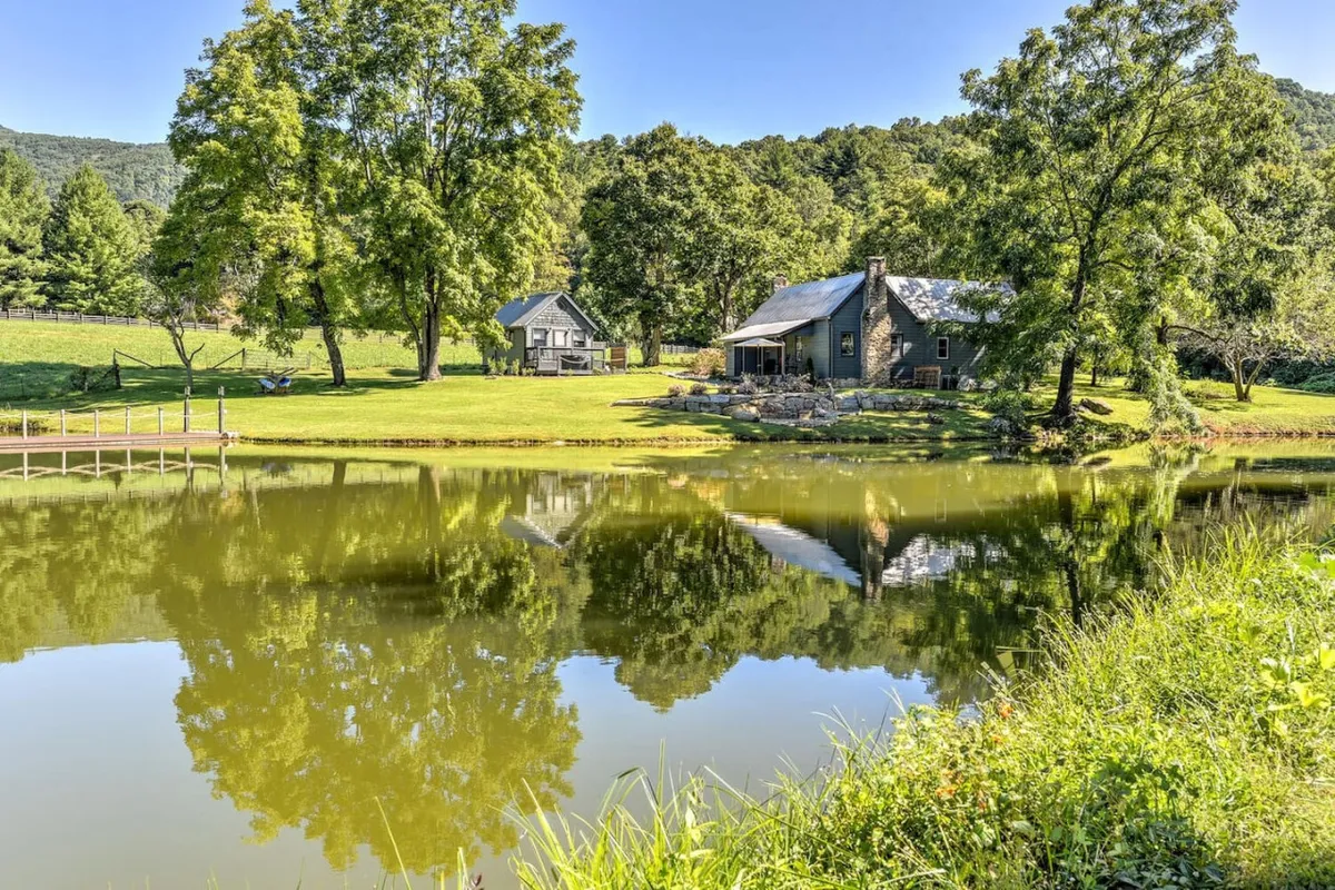 The hero image features a tranquil scene with a picturesque pond reflecting the soft hues of sunset, surrounded by lush trees. The charming, newly renovated farmhouse and its guest cottage are nestled beside the water, creating an inviting and peaceful atmosphere. The wide, open grounds suggest ample space for fishing, picnicking, and relaxing by the water's edge.