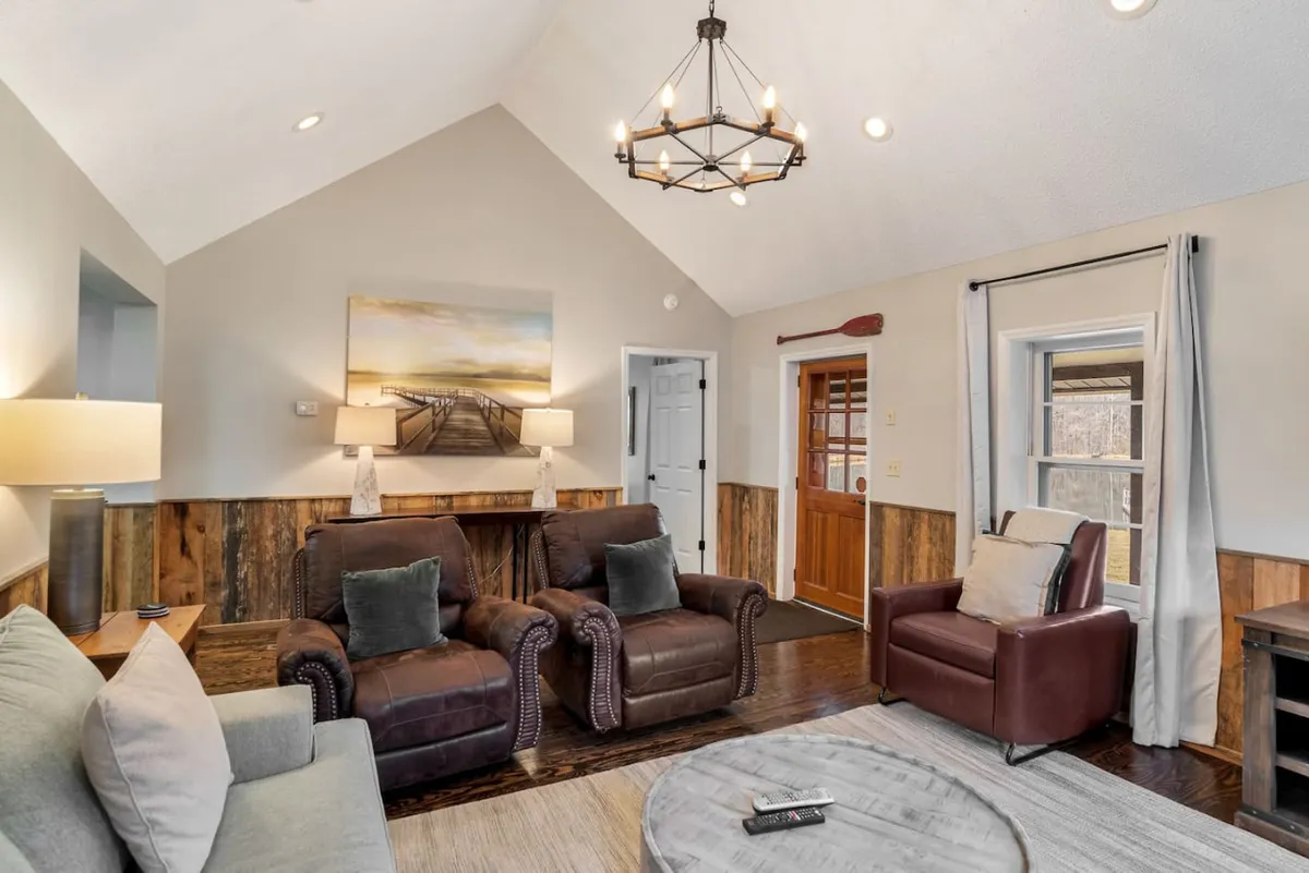 A welcoming space highlighted by a striking chandelier, large artwork, and comfortable leather armchairs situated near a wooden entrance door. Generous windows allow natural light to flood the room, reflecting off the wood floors and creating a bright, open feel ideal for casual conversations or morning coffee.