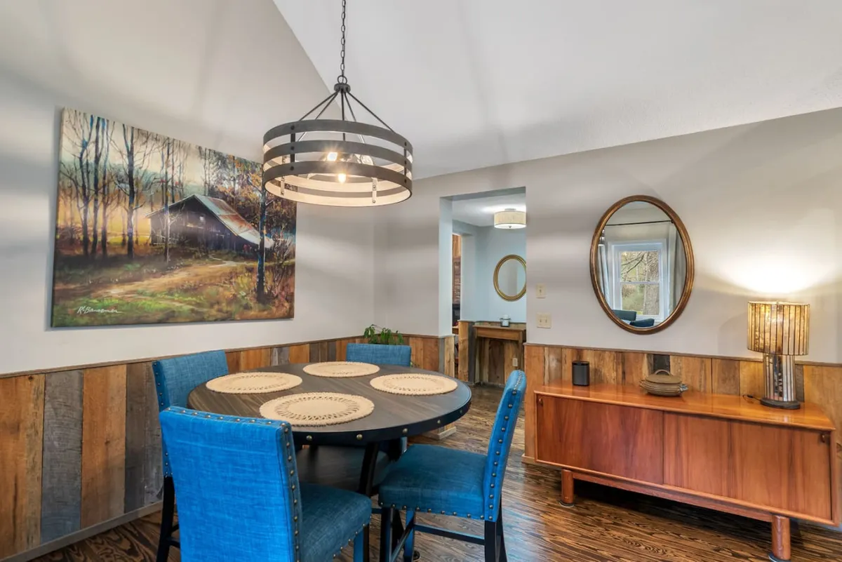 A round dining table surrounded by stylish blue upholstered chairs sits beneath a modern light fixture. Artwork and a wooden sideboard add character, while natural light from windows enhances the warm, welcoming ambiance—ideal for intimate meals or casual gatherings.
